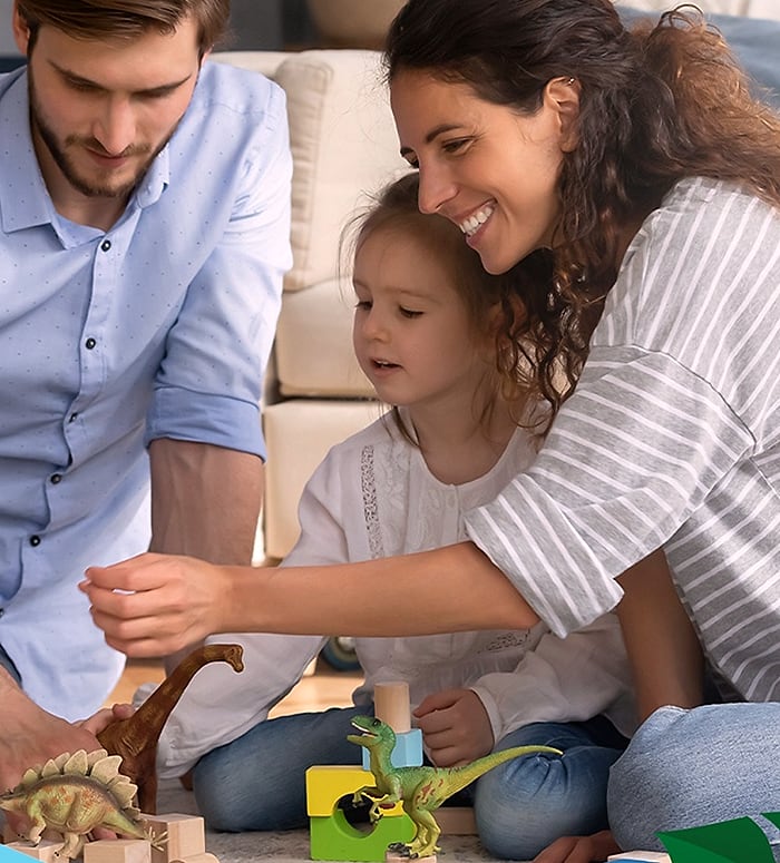 Parents and a young child playing with toy dinosaurs and wooden blocks on the floor
