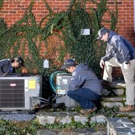 Technicians working on an outdoor heat pump unit beside a brick house